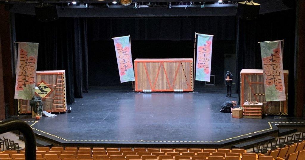 Wide view of a theater stage at The Reif set up for a dinosaur-themed production, featuring large crate props, colorful ‘Dinosaur World’ banners, and crew members preparing the stage before a performance.