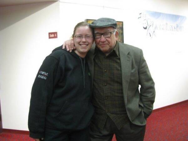 Two people smile and pose together in a theater or event lobby, with one person wearing glasses and a black hoodie and the other wearing a flat cap, glasses, and a blazer.