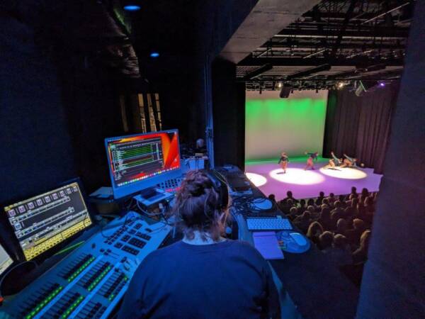 “Lighting technician works at a control booth overlooking a live dance performance at The Reif, with stage lighting equipment and computer monitors visible beside the audience and performers on stage below.