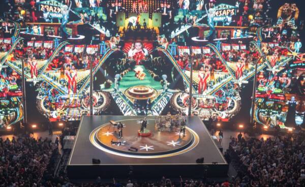 Wide view of a live arena concert featuring a band performing on a circular stage surrounded by a large crowd, with towering colorful video screens displaying vibrant collage-style visuals behind the performers.