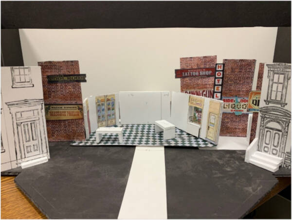 Detailed scale model of a theater set featuring brick city storefronts, a hotel sign, and a partially opened interior room with tiled flooring and miniature stage platforms arranged along a street scene.
