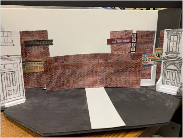 Scale model of an urban street theater set featuring brick building facades, storefront signs for a tattoo shop and hotel, and miniature architectural details arranged on a stage platform.