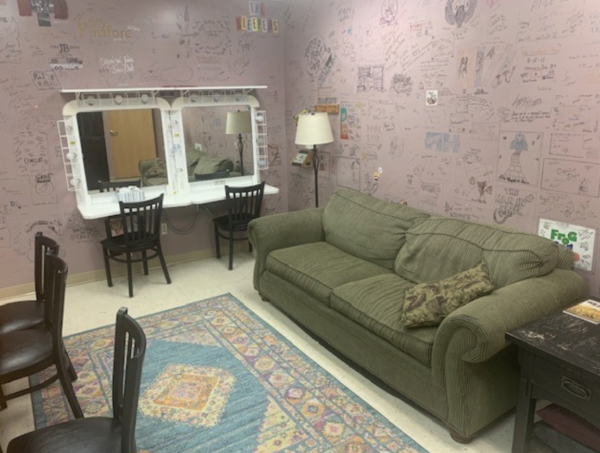 Cozy backstage dressing room at The Reif featuring a green couch, vanity mirrors with lights, chairs, a patterned rug, and walls covered with handwritten signatures, doodles, and messages from performers.