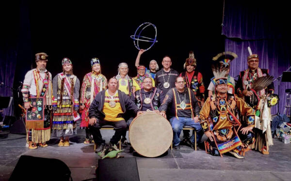 Group portrait of Indigenous performers and drummers in colorful traditional regalia gathered on a theater stage, posing with a large drum and ceremonial items during a cultural performance at The Reif.