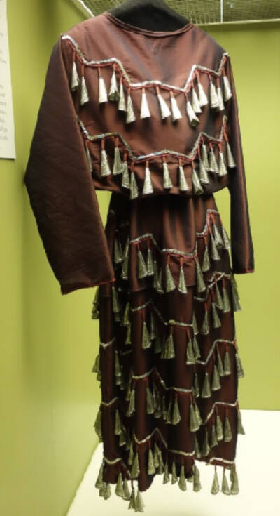 Brown theatrical costume displayed on a mannequin, featuring layered rows of decorative tassels across the sleeves and skirt, hanging against a light green museum or costume archive wall.