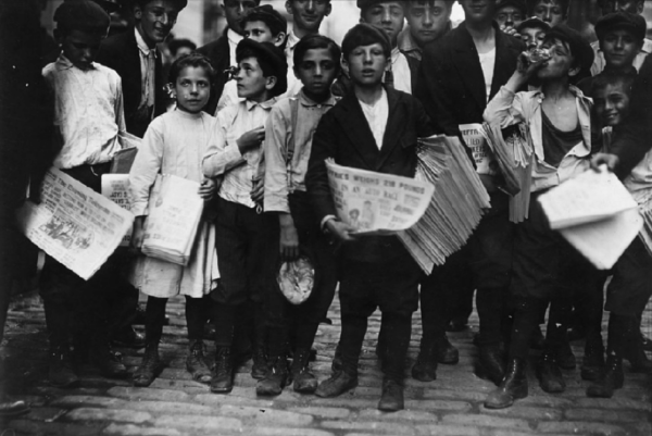Historic black-and-white photograph of young newsboys and children standing together on a cobblestone street holding stacks of newspapers, with adults gathered behind them in an early 1900s city scene.
