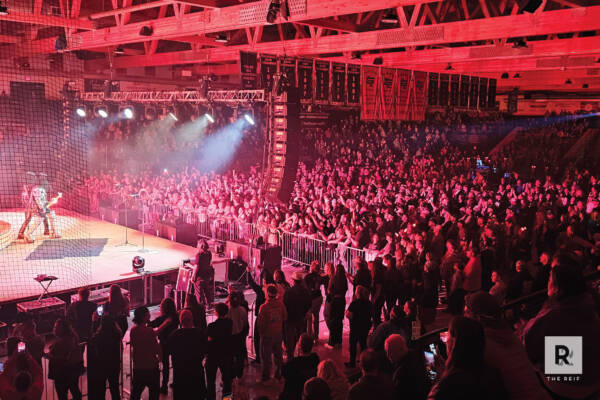 Wide view of a packed concert crowd inside an arena-style venue at The Reif, illuminated by dramatic red stage lighting as a performer plays on stage behind security barriers and overhead truss lighting.