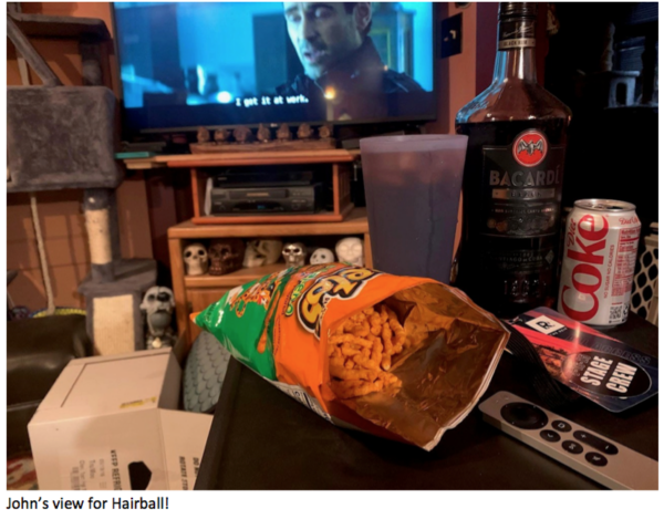 Casual living room setup with an open bag of Cheetos, a bottle of Bacardi, a Diet Coke can, and a TV remote on a table while a movie or TV show plays in the background, with skull decorations displayed beneath the television and the caption ‘John’s view for Hairball!!’ at the bottom.