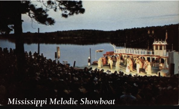 Outdoor nighttime performance of Mississippi Melodie Showboat staged beside a lake, featuring actors performing on a large riverboat-style set while a seated audience watches from a hillside amphitheater. Text in the corner reads: ‘Mississippi Melodie Showboat.