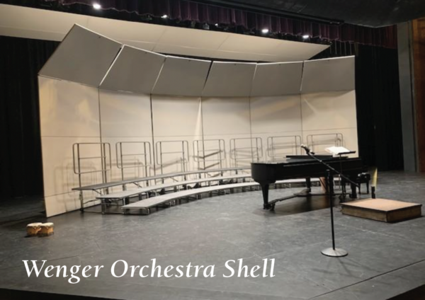Concert choir setup on a theater stage featuring a curved Wenger orchestra shell, tiered choral risers, a grand piano, microphone stand, and conductor’s podium beneath stage lighting. Text in the lower corner reads: ‘Wenger Orchestra Shell.