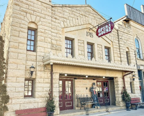 Historic stone-front opera house building with a vintage marquee sign reading ‘Opera House’ and the year ‘1918’ displayed above the entrance. The restored facade features tall windows, decorative trim, and double front doors along a small-town street.