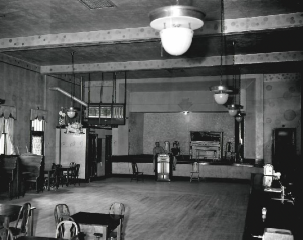Historic black-and-white photograph of a large early 20th-century dance hall or ballroom interior with a wooden floor, hanging globe lights, scattered tables and chairs, a balcony, and a small stage area with people standing near a piano in the background.