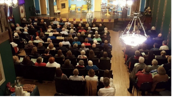 View from a balcony overlooking a packed theater audience watching a live stage performance in an elegant historic-style auditorium with chandeliers, wood floors, and a brightly lit set featuring living room furniture.
