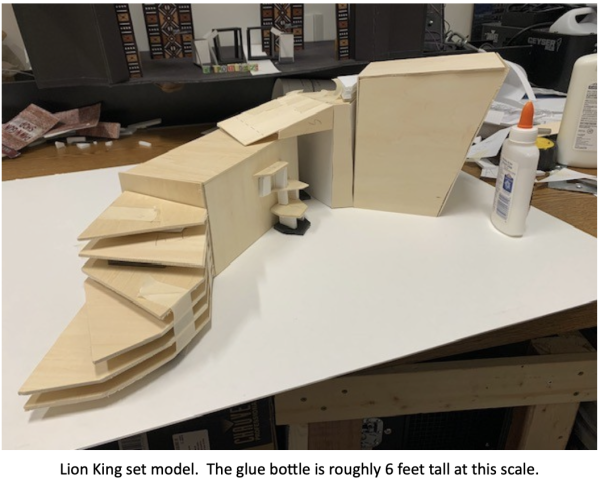 Wooden scale model for a Lion King stage set displayed on a worktable, featuring layered steps, platforms, and an angular Pride Rock structure beside a bottle of glue used to show scale. Caption at the bottom reads: ‘Lion King set model. The glue bottle is roughly 6 feet tall at this scale.