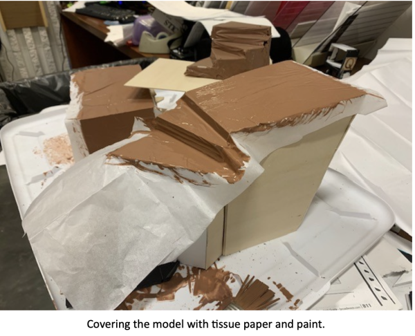 Behind-the-scenes model-building process showing a miniature scenic structure covered with tissue paper and brown paint to create textured terrain or rock surfaces, with paint drips and craft materials scattered around the workspace. Caption at the bottom reads: ‘Covering the model with tissue paper and paint.