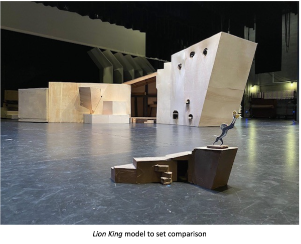 Scale comparison photograph for a Lion King stage set design showing large angular scenic structures and platforms on a theater stage, with a small miniature human figure model in the foreground for size reference. Caption at the bottom reads: ‘Lion King model to set comparison.