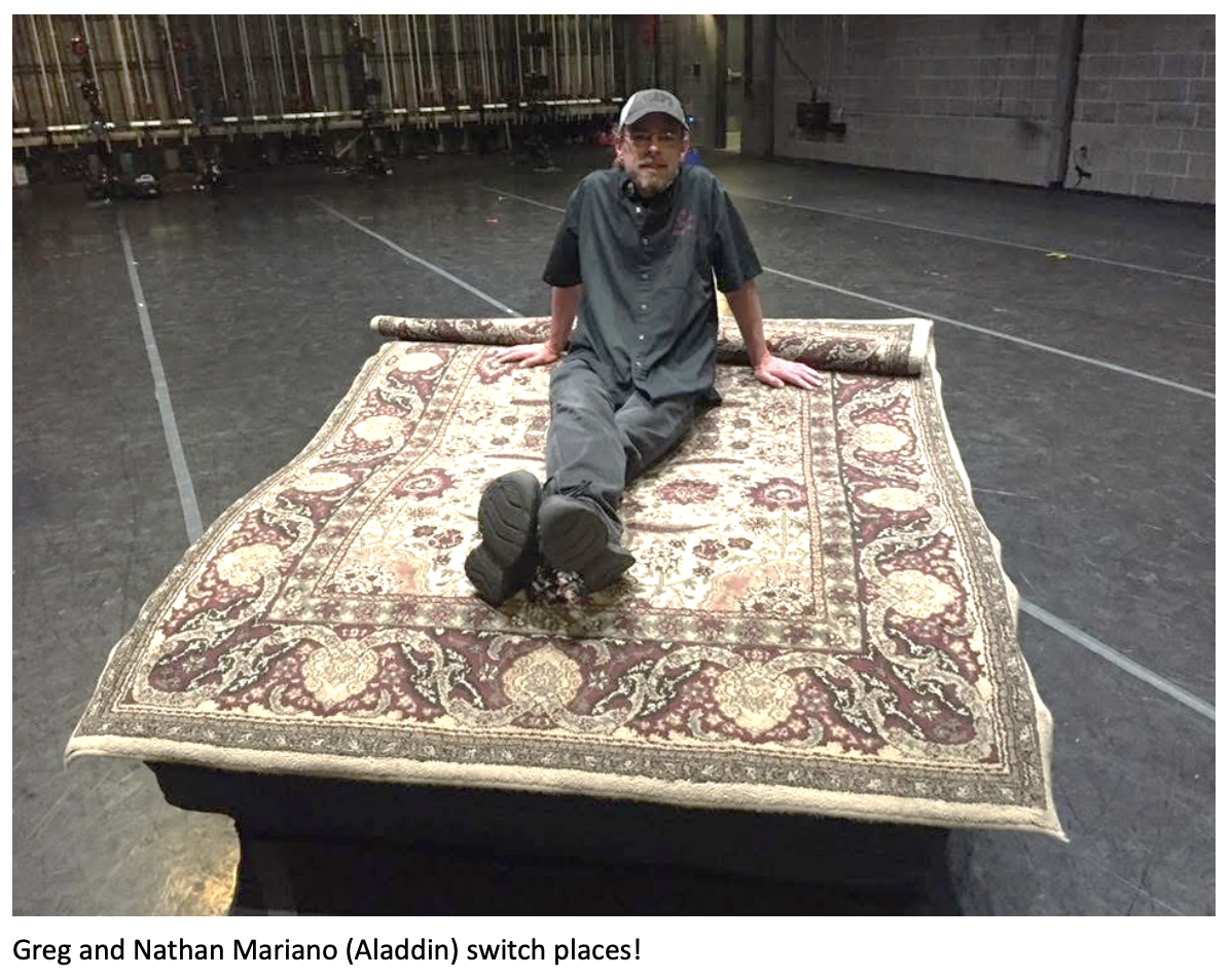 Man wearing a cap and dark work clothes sits on a large patterned rug prop placed on a theater stage, humorously recreating a flying carpet pose from Aladdin. Caption at the bottom reads: ‘Greg and Nathan Mariano (Aladdin) switch places!’