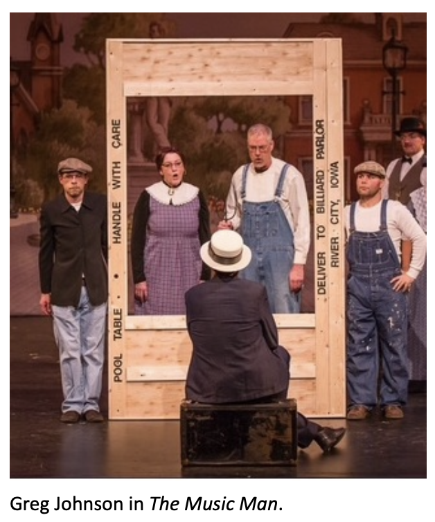 Stage scene from The Music Man featuring several performers gathered around an oversized wooden shipping crate standing upright like a giant frame. A seated performer in a straw boater hat faces the crate while actors in period costumes and overalls pose around it on a painted town backdrop stage set. Caption at the bottom reads: ‘Greg Johnson in The Music Man.