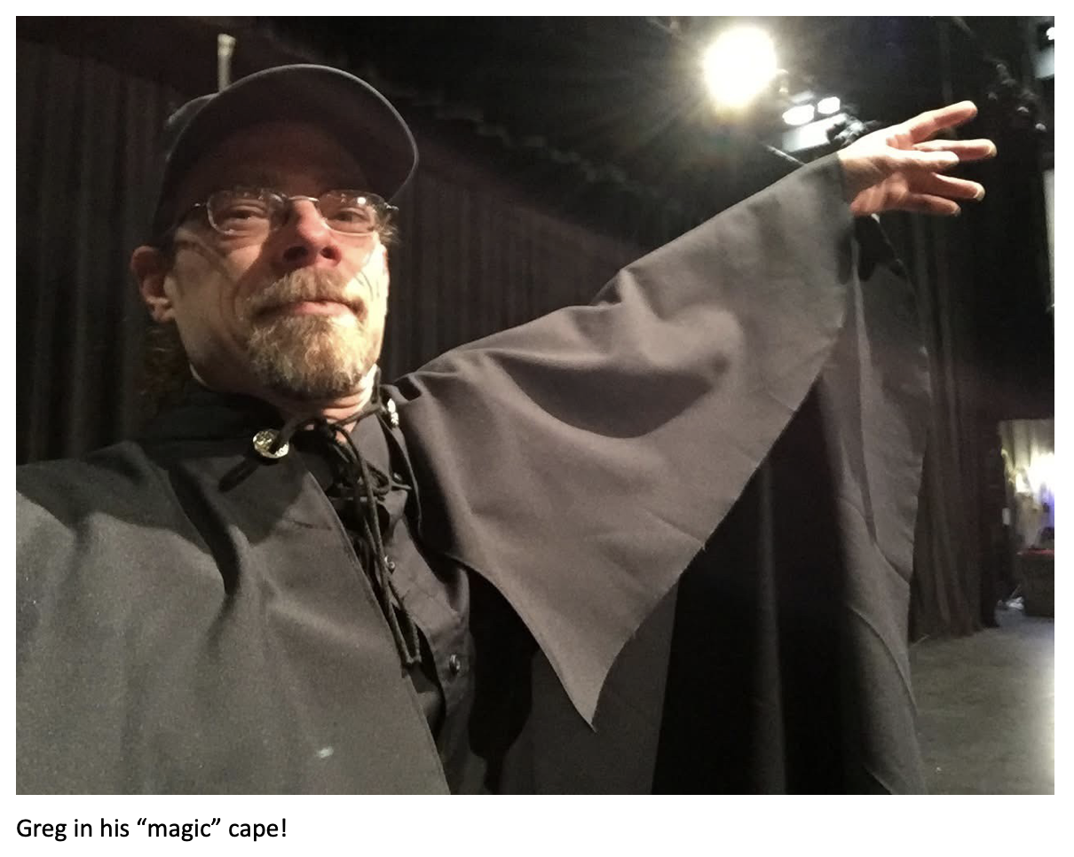 Man wearing glasses, a baseball cap, and a dramatic black cape poses backstage on a theater stage with one arm extended theatrically under bright stage lights. Caption at the bottom reads: ‘Greg in his “magic” cape!