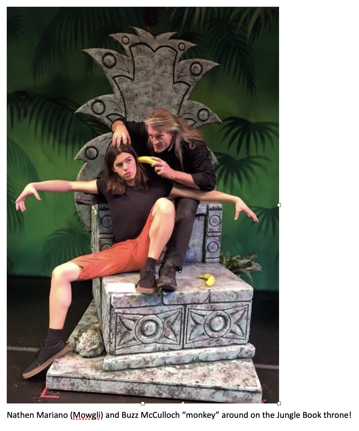 Two performers pose playfully on a large stone jungle throne prop during a rehearsal or production of The Jungle Book. One performer lounges dramatically across the throne while the other leans over mischievously holding a banana, with tropical foliage painted on the backdrop behind them. Caption at the bottom reads: ‘Nathen Mariano (Mowgli) and Buzz McCulloch “monkey” around on the Jungle Book throne!