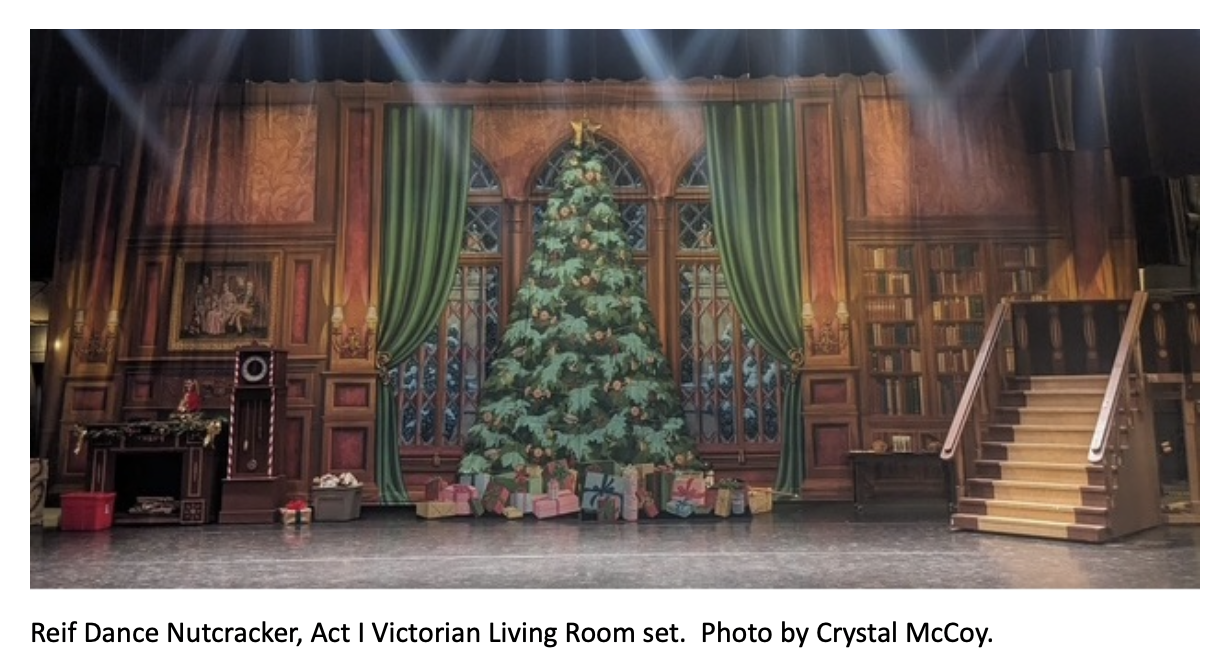 Elaborate Victorian-style living room stage set for The Nutcracker, featuring a towering decorated Christmas tree at center stage, wrapped presents underneath, tall arched windows with green curtains, bookshelves, a fireplace, and a staircase leading upward on the right side under theatrical lighting. Caption at the bottom reads: ‘Reif Dance Nutcracker, Act I Victorian Living Room set. Photo by Crystal McCoy.