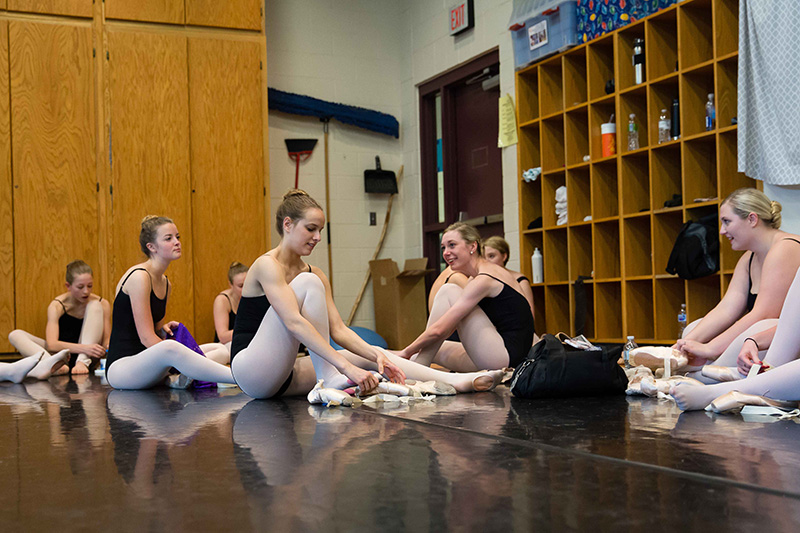 Ballet students in black leotards and white tights sit on the studio floor preparing and adjusting their pointe shoes during class.