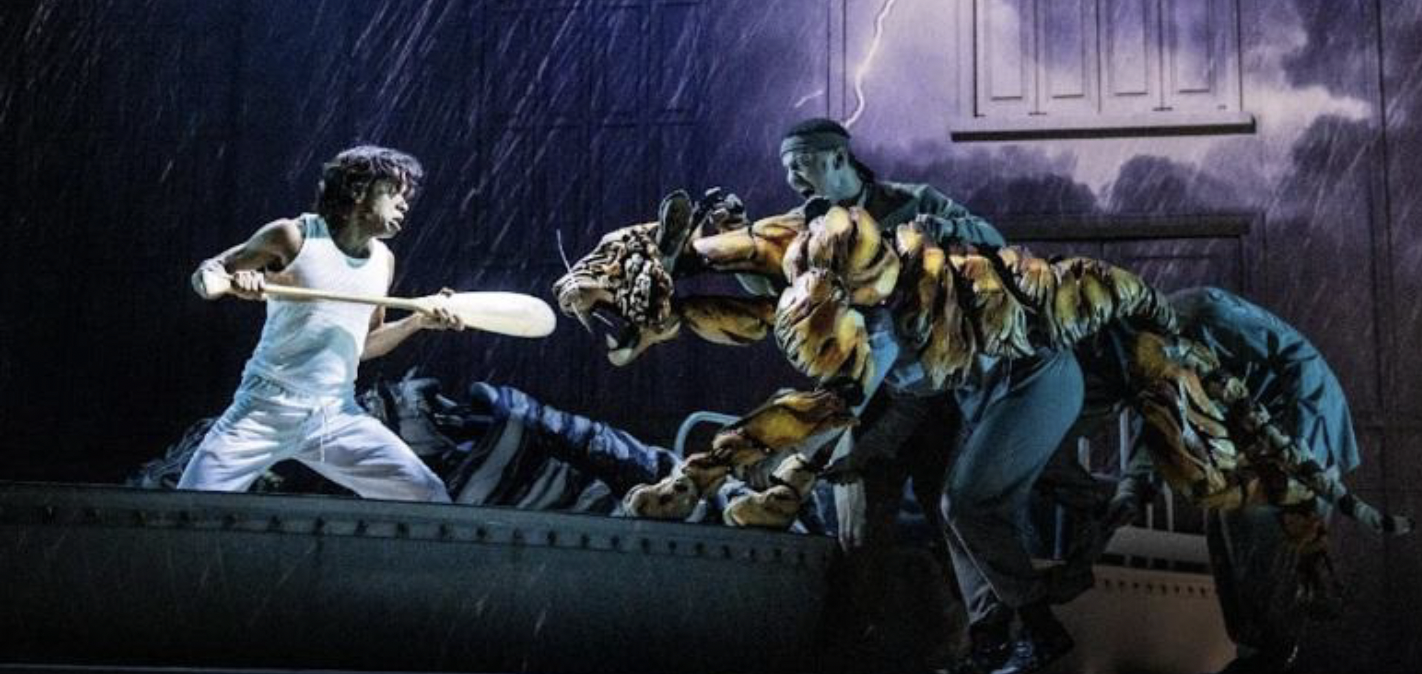 A dramatic stage scene shows a young performer in white pajamas holding a wooden paddle defensively while confronting a large tiger puppet operated by performers dressed in dark clothing. Rain and lightning effects create a tense nighttime atmosphere onstage.