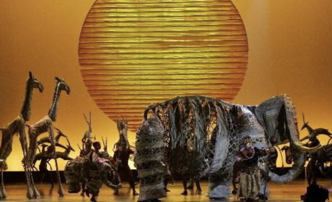 A stage performance inspired by The Lion King features large animal puppets and performers silhouetted against a glowing orange backdrop with a giant sun. A life-sized elephant puppet dominates center stage, surrounded by giraffes, antelope figures, and dancers in stylized costumes.