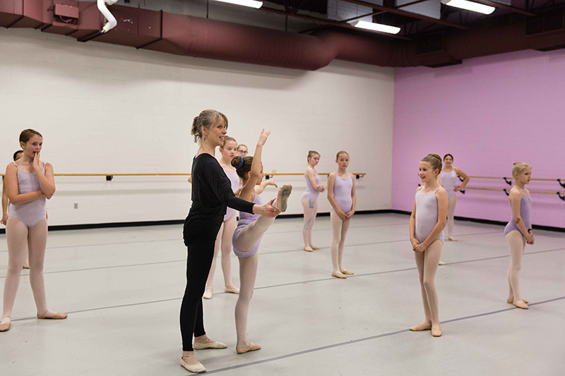 A ballet instructor assists a young student with a leg stretch in a bright dance studio while other children in lavender leotards watch and practice nearby.