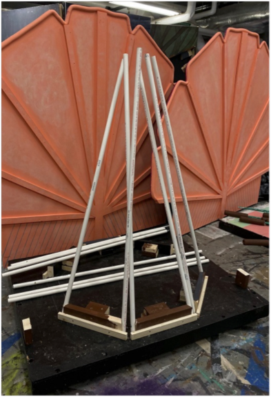 Partially constructed stage prop stands in a theater workshop, featuring a pyramid-shaped frame made from white PVC pipes mounted on a black platform, with large orange decorative panels in the background.