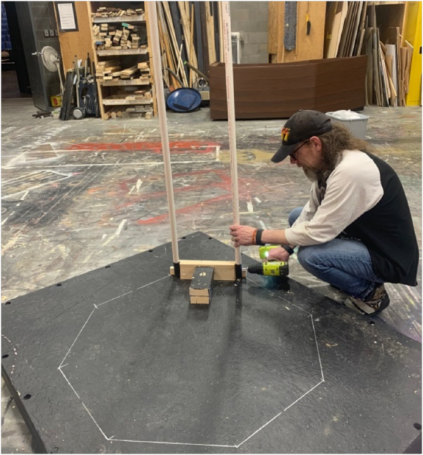 Person works in The Reif’s scene shop using a power drill to assemble a stage prop frame made from white PVC pipes on a black platform marked with chalk guidelines, surrounded by woodworking materials and theater set pieces.