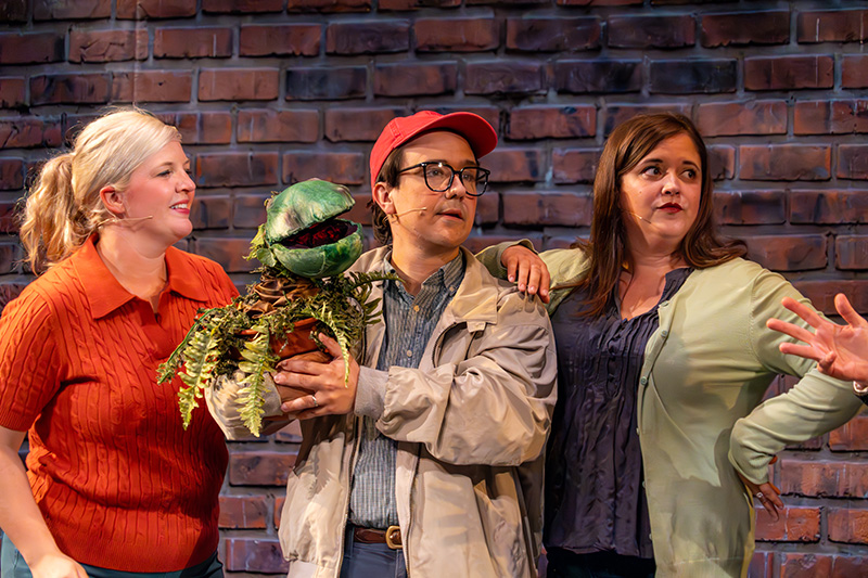 Actors perform a musical theater scene on stage at The Reif, featuring a performer holding a green plant puppet while standing between two cast members in front of a brick wall set.