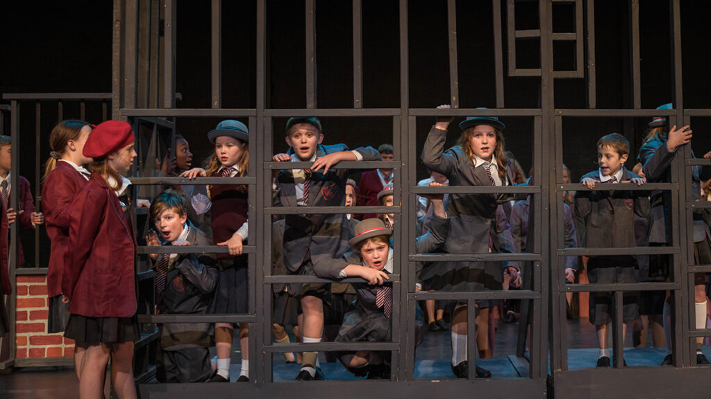 Children in school uniforms perform on stage at The Reif during a youth theater production, standing among large metal framework set pieces under theatrical lighting.