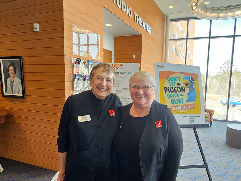 Two volunteers smile in the lobby of The Reif near the Studio Theater entrance, standing beside a poster for ‘Don’t Let the Pigeon Drive the Bus! The Musical!’