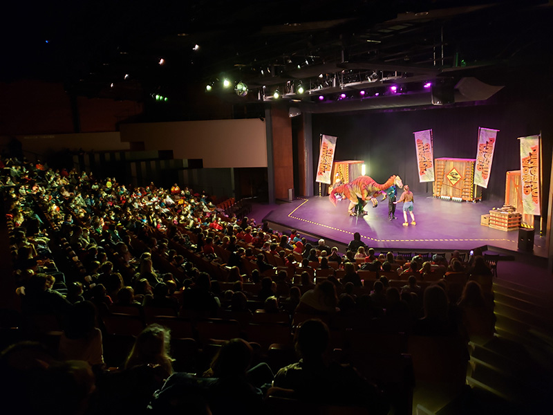 A large audience fills the auditorium at the Reif Center while performers present a live dinosaur-themed stage show under colorful theatrical lighting. A dinosaur puppet stands center stage beside performers, with themed banners and set pieces in the background.