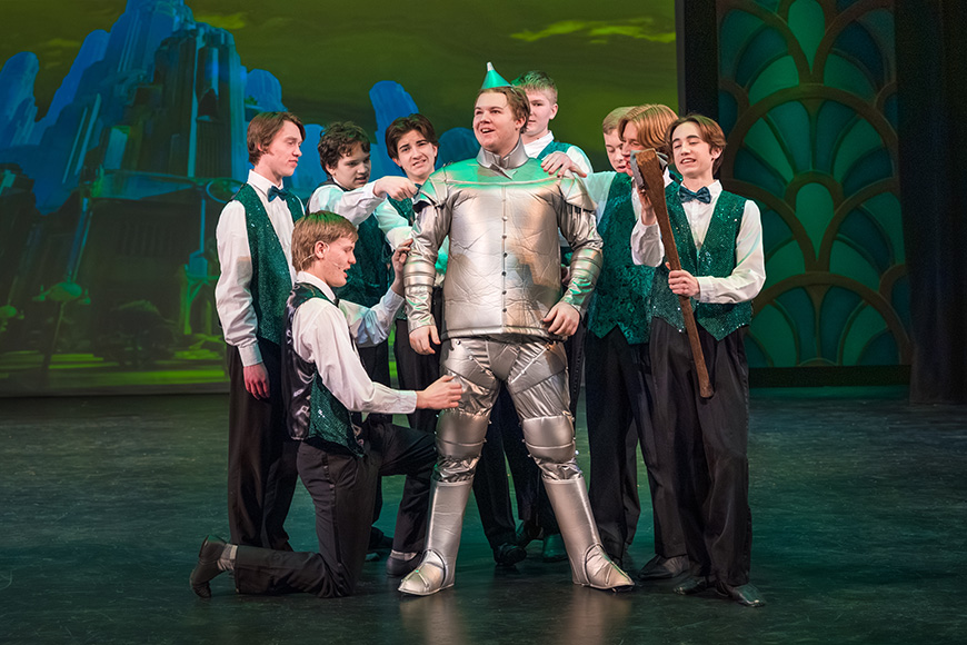 A group of young performers gathers around a student dressed as the Tin Man during a musical theater production at The Reif, with colorful stage lighting and an Emerald City-inspired backdrop.