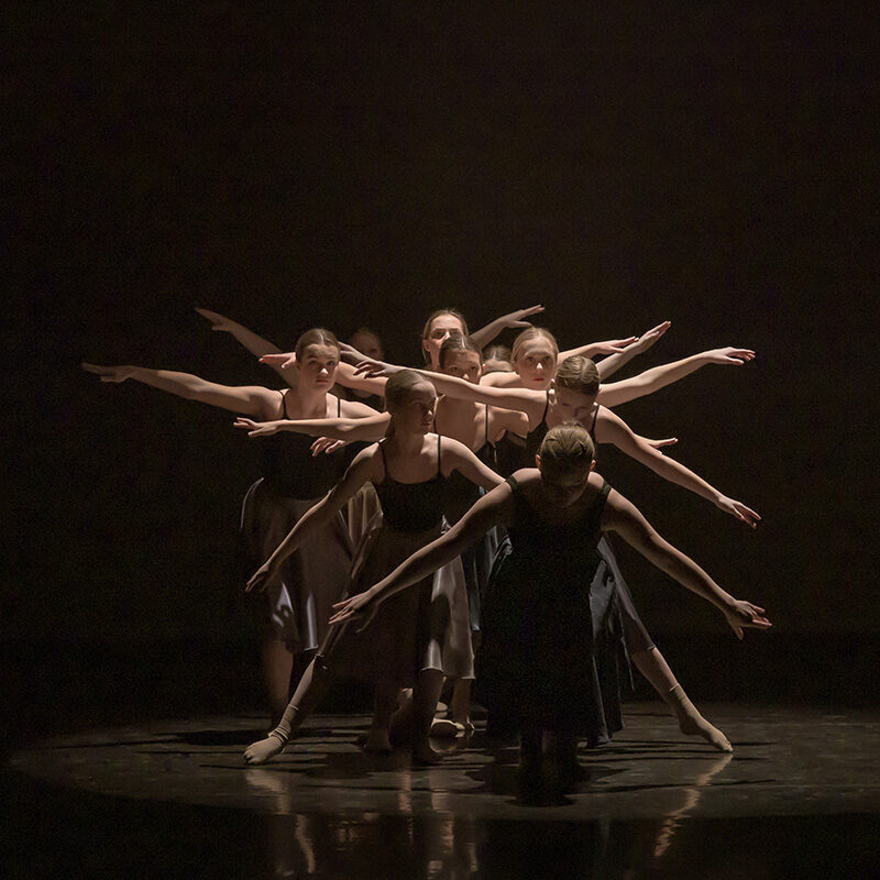 A group of young dancers performs on stage at The Reif under dramatic spotlighting, creating a layered formation with extended arms against a dark background